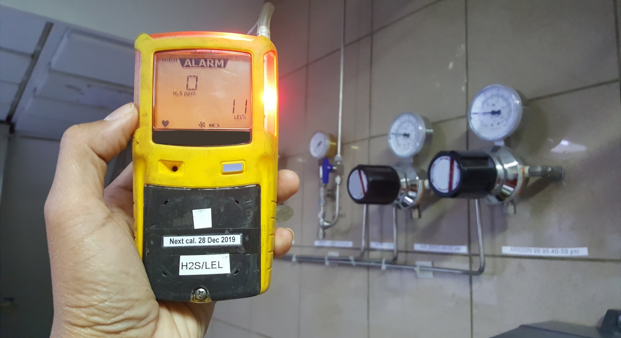 A hand holding a yellow digital gas detector displaying an alarm, with industrial pressure gauges and piping blurred in the background.
