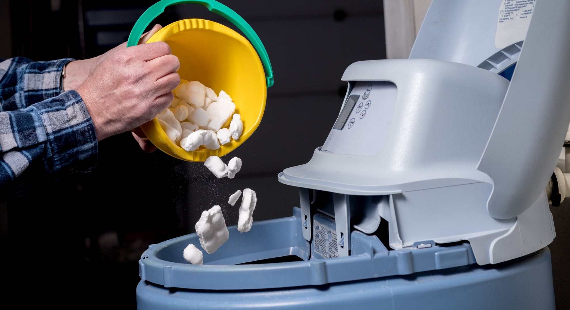 A person pours white salt blocks from a small yellow bucket into the brine tank of a water softener system.