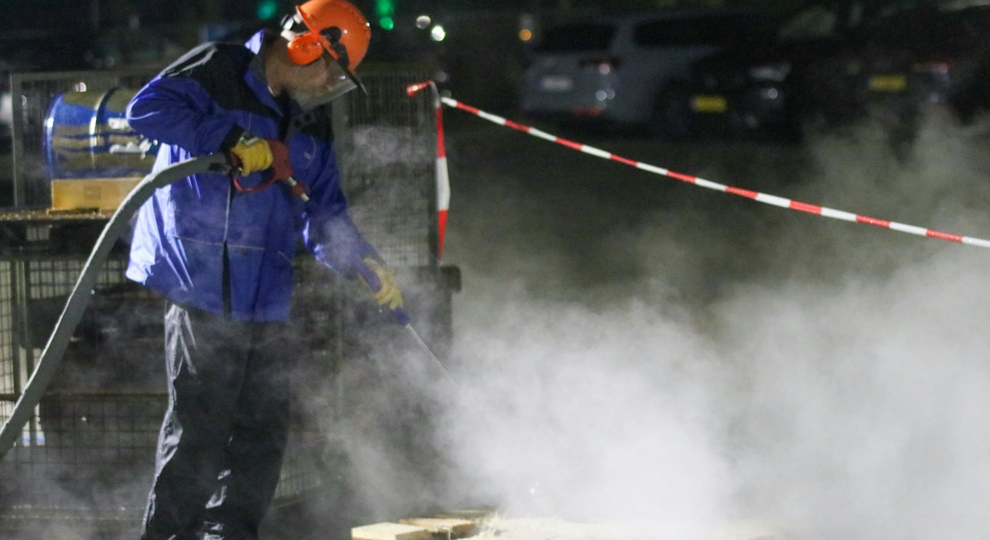 A worker wearing full safety gear and an orange helmet uses a high-pressure water jetting hose at night, surrounded by steam.