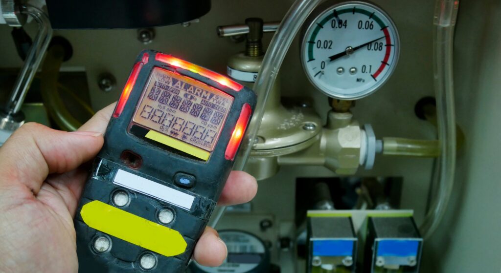 A close-up of a hand holding a digital gas detector with a lit red alarm indicator, positioned in front of a pressure gauge and industrial machinery.