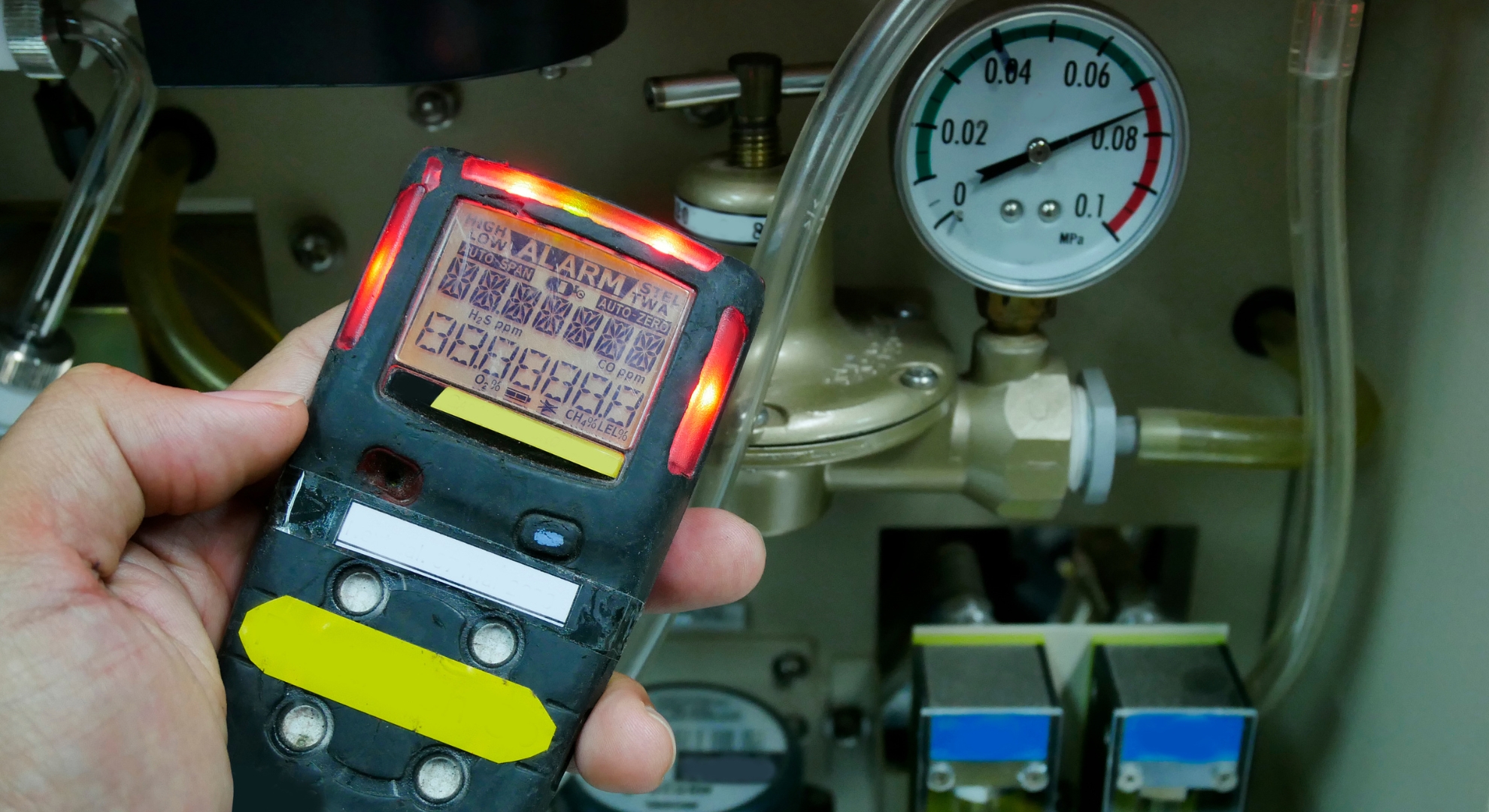 A close-up of a hand holding a digital gas detector with a lit red alarm indicator, positioned in front of a pressure gauge and industrial machinery.