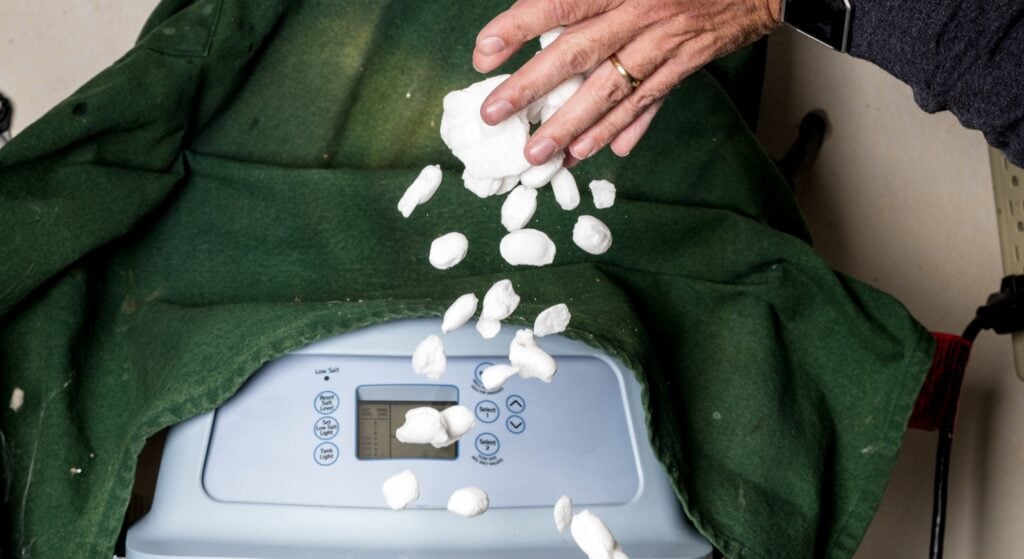 A hand dropping white salt pellets into the top opening of a water softener system.