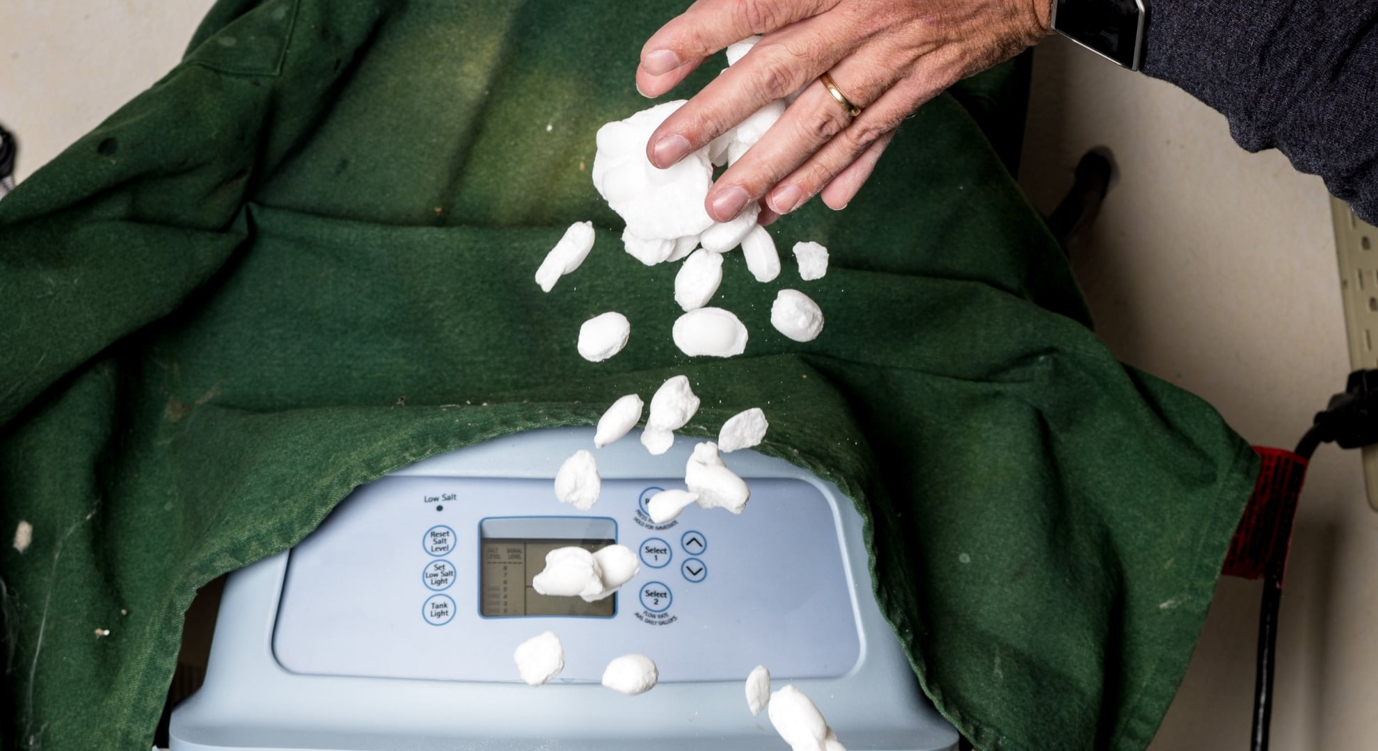 A hand dropping white salt pellets into the top opening of a water softener system.