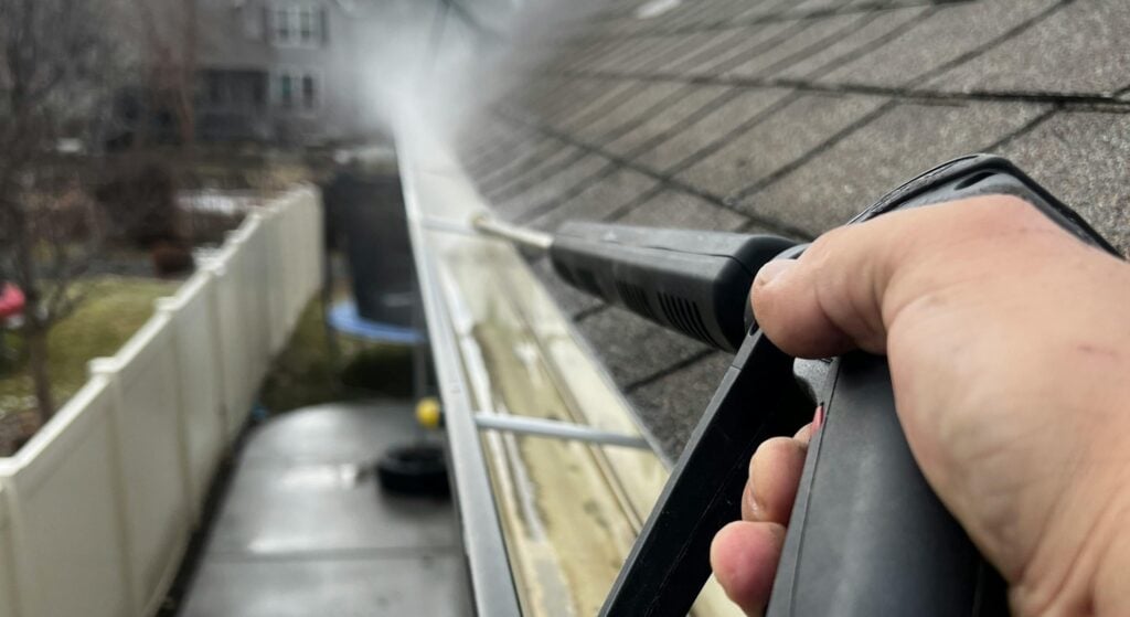 First-person view of a person using a high-pressure water sprayer to clean a roof gutter.
