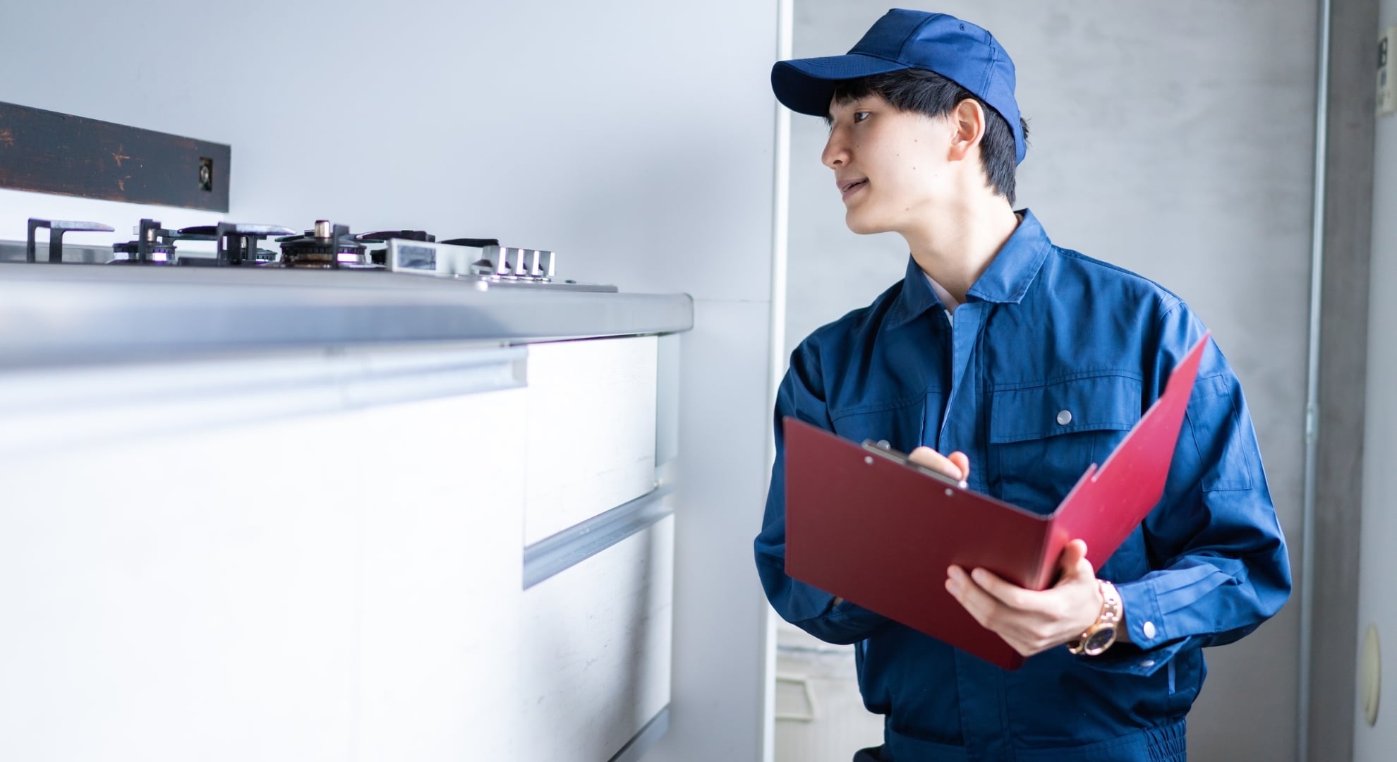 A technician wearing a blue uniform and cap looks at a kitchen gas stovetop while writing on a clipboard.